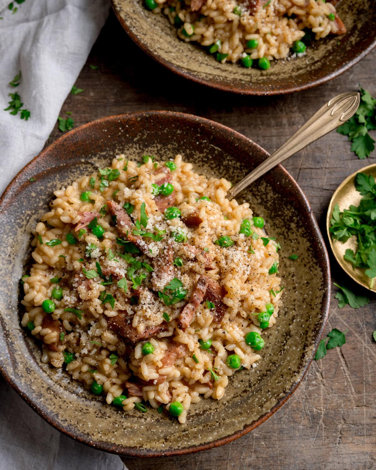 Ham and Pea Risotto served in a rustic brown bowl with a fork sticking out. The bowl is on a wooden table and there is a further bowl nearby, as well as a light napkin and a small gold plate with chopped parsley on it.