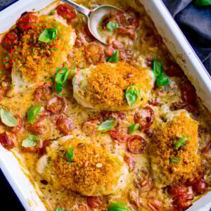 A white baking dish with four breaded chicken breasts in a creamy pesto sauce with roasted cherry tomatoes and basil leaves. There is a spoon resting in the top of the dish.