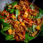 Pineapple pork stir-fry in a wok with pork strips, crisp mangetout, pineapple chunks, red peppers, and chillies in a savoury sauce topped with chopped spring onions.