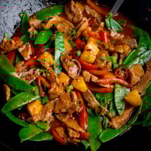 Pineapple pork stir-fry in a wok with pork strips, crisp mangetout, pineapple chunks, red peppers, and chillies in a savoury sauce topped with chopped spring onions.