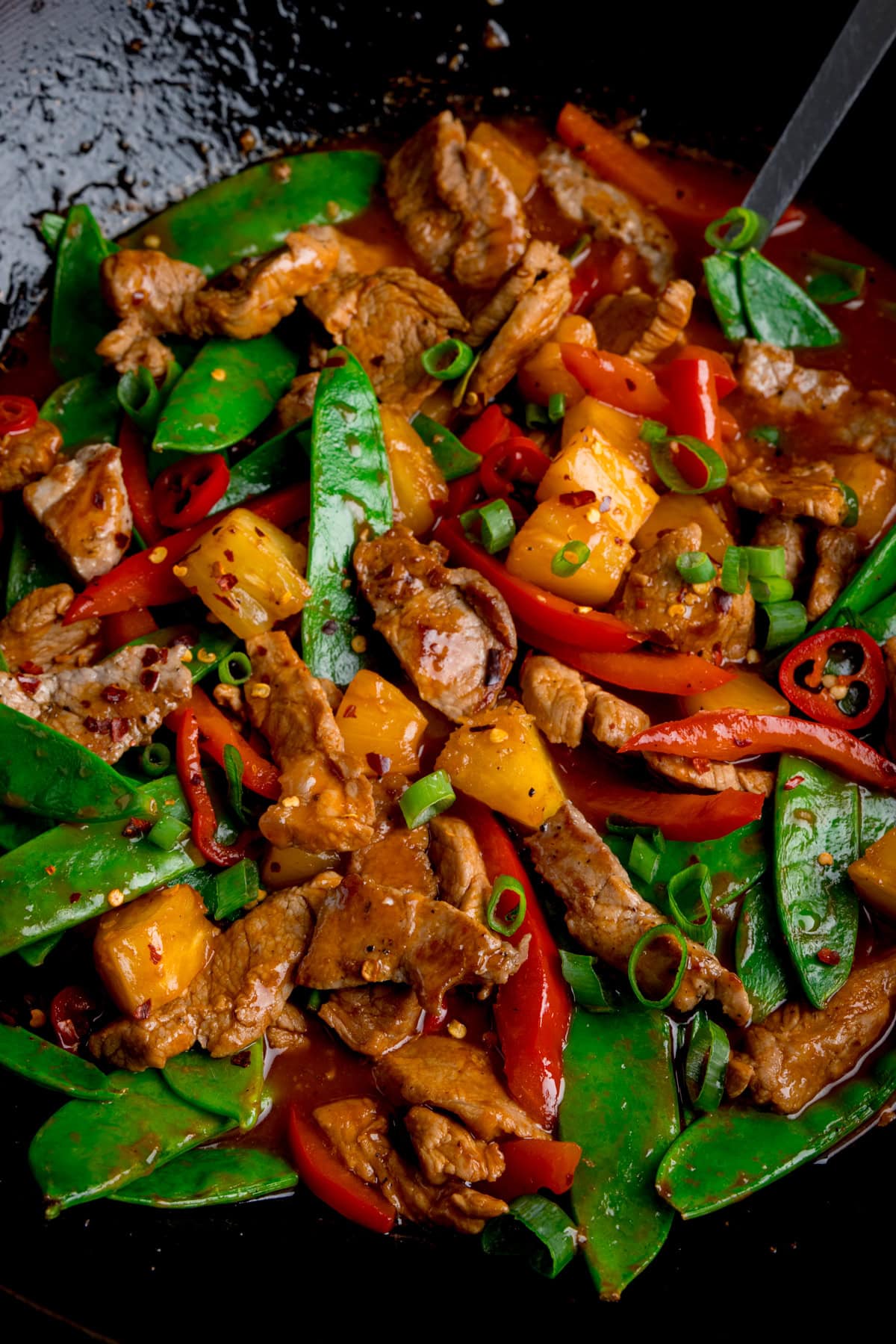 Stir-fry with sliced pork, mangetout, red peppers, pineapple pieces, and spring onions in a glossy sauce, served in a black wok.