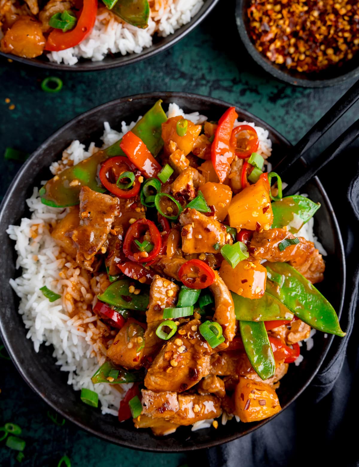 A bowl of white rice topped with pineapple pork stir fry with mangetout, pineapple, and spring onions, served with a black chopsticks.