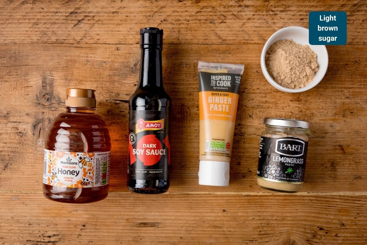 Five ingredients on a wooden surface: a bottle of honey, dark soy sauce, ginger purée, lemongrass purée, and a bowl of light brown sugar.