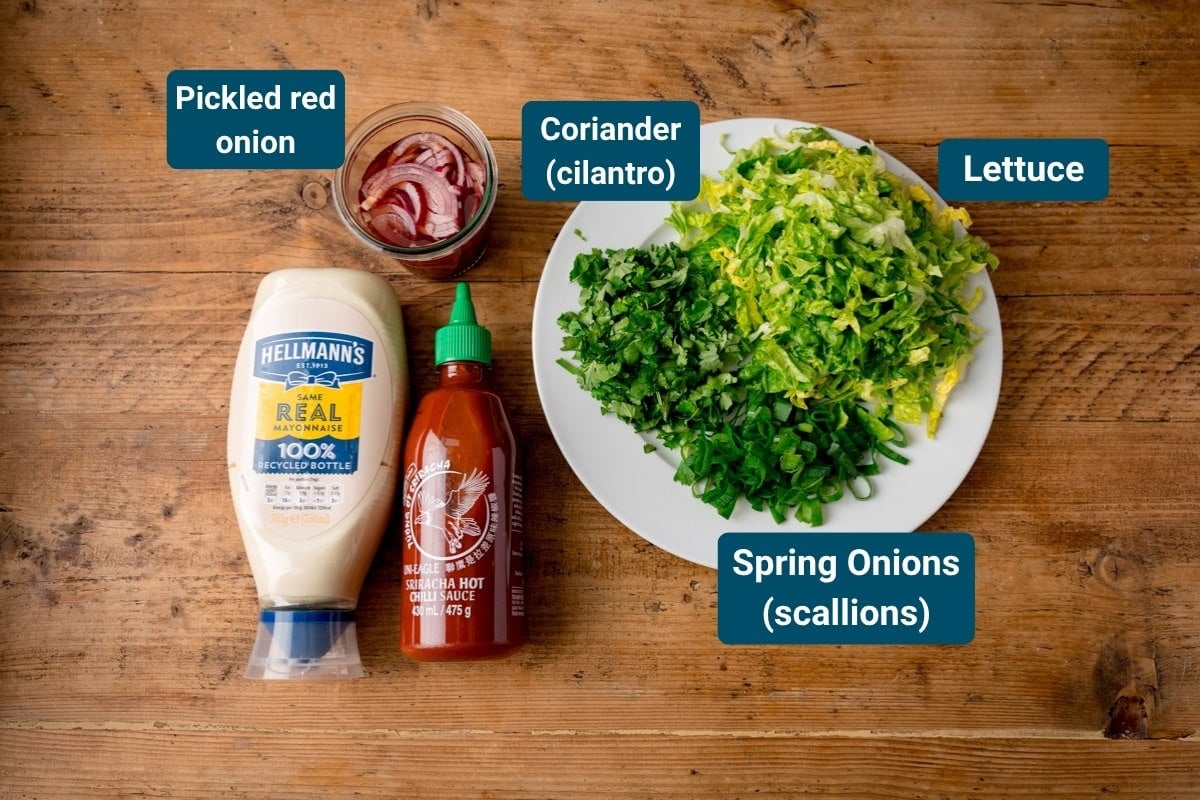A plate with chopped lettuce, coriander, and spring onions sits next to a jar of pickled red onion, mayonnaise, and sriracha sauce on a wooden table.
