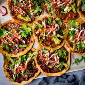 Overhead image of pork smash tacos topped with shredded lettuce, pickled onions, coriander, mayonnaise, and sriracha on a silver baking tray with chopped spring onions.
