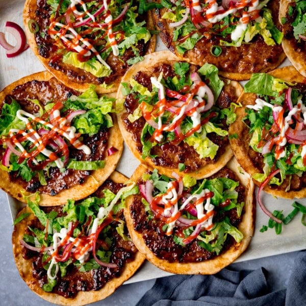 Overhead image of pork smash tacos topped with shredded lettuce, pickled onions, coriander, mayonnaise, and sriracha on a silver baking tray with chopped spring onions.