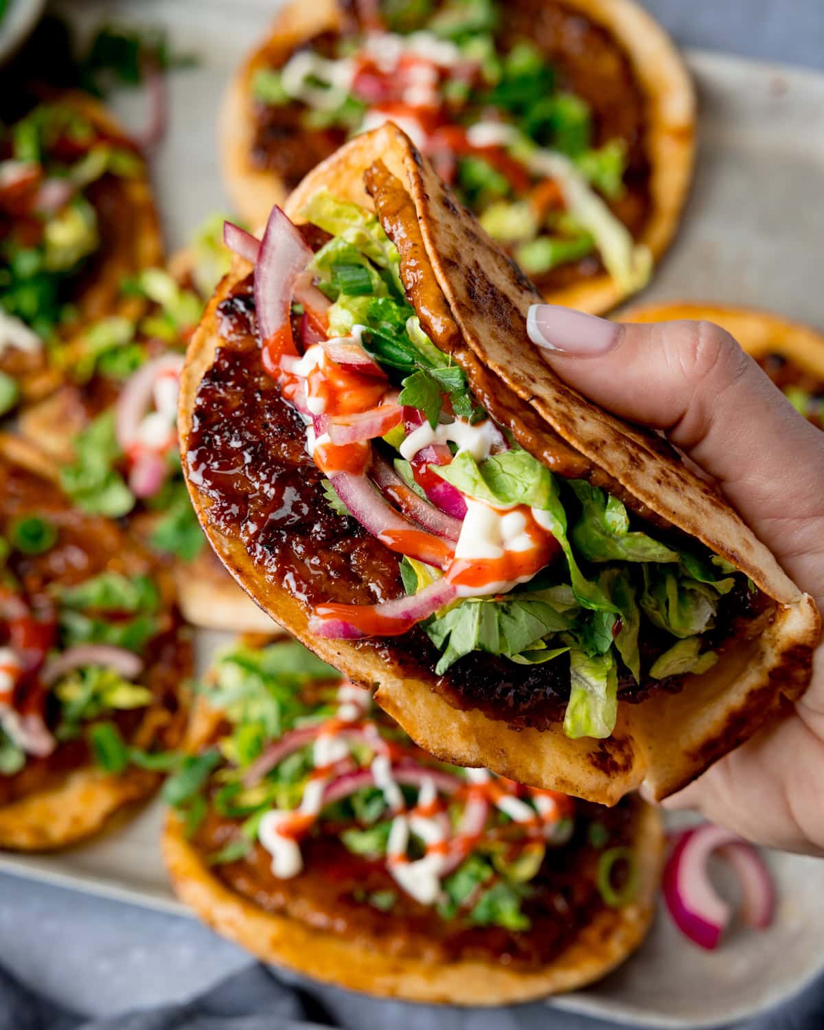 A hand holds a folder pork smash taco topped with lettuce, sliced red onions and sauce. Several further smash tacos are on a tray in the background.