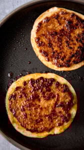 Two smash tacos are cooking in a black frying pan. The meat is cooked and golden.