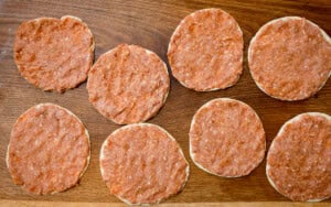 Eight soft tacos topped with seasoned ground pork that has been flattened to fit the size of the tacos. The uncooked smash tacos are on a wooden surface.