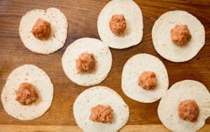 Eight soft tacos, each topped with a ball of raw, seasoned ground pork, ready to turn into smash tacos. The tacos are on a wooden surface.