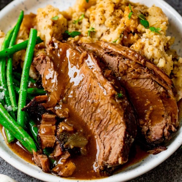 Sliced slow cooker roast beef with brown gravy, served with mashed potato and green beans on a white plate.