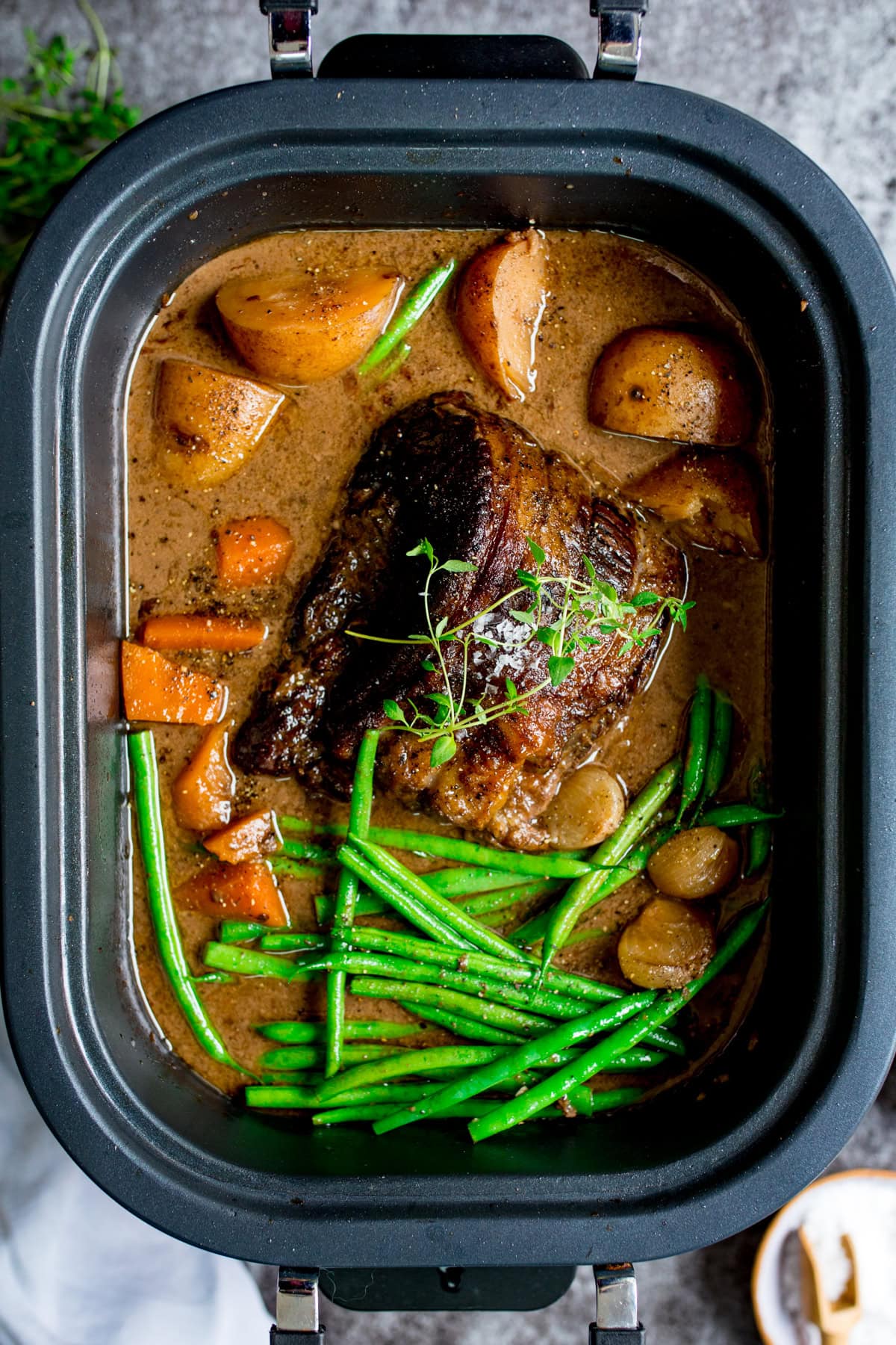 A cooked pot roast with gravy, potatoes, carrots, green beans, and herbs in a black slow cooker.
