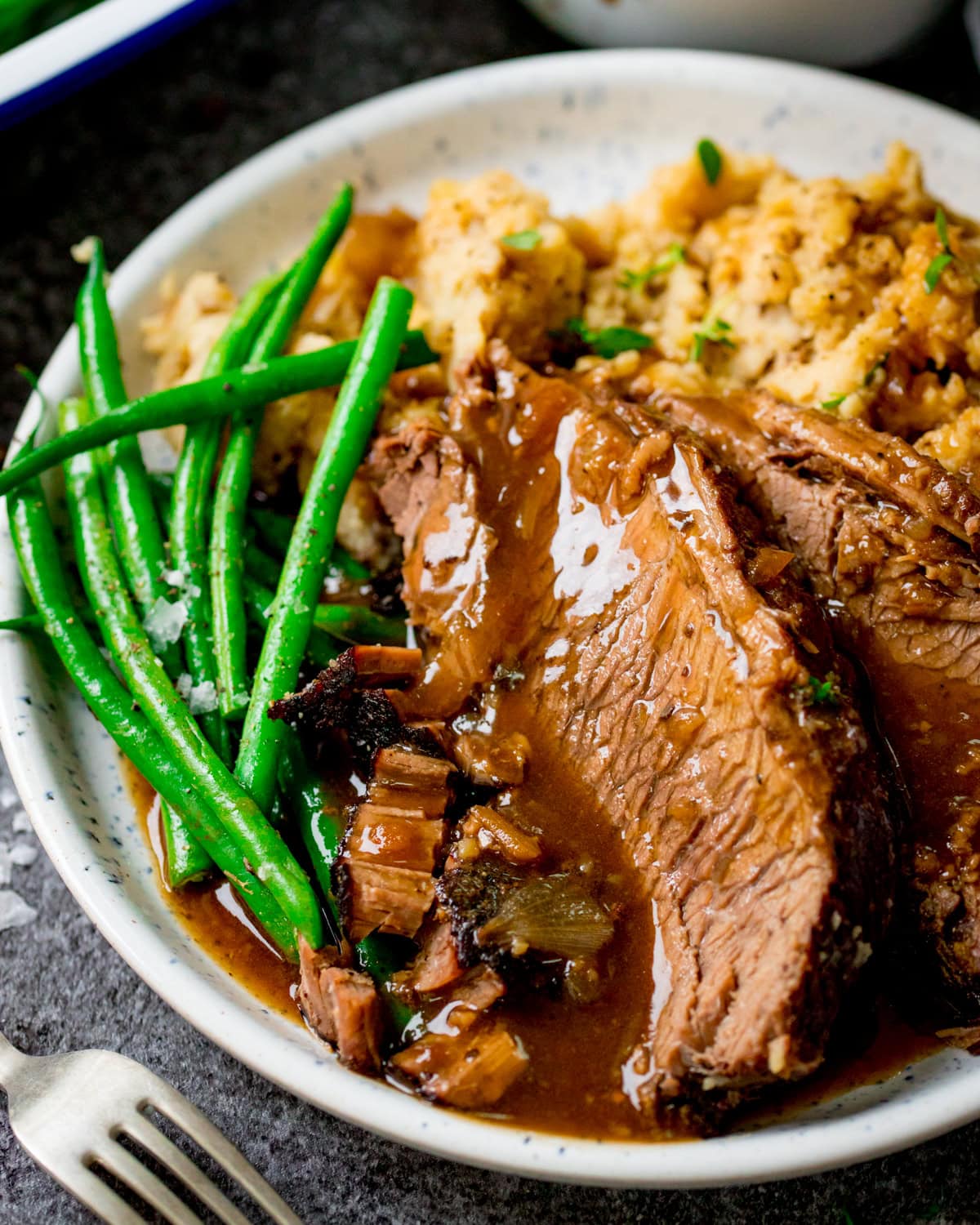 A plate with slices of crockpot roast beef in brown gravy, green beans, and mashed potato.