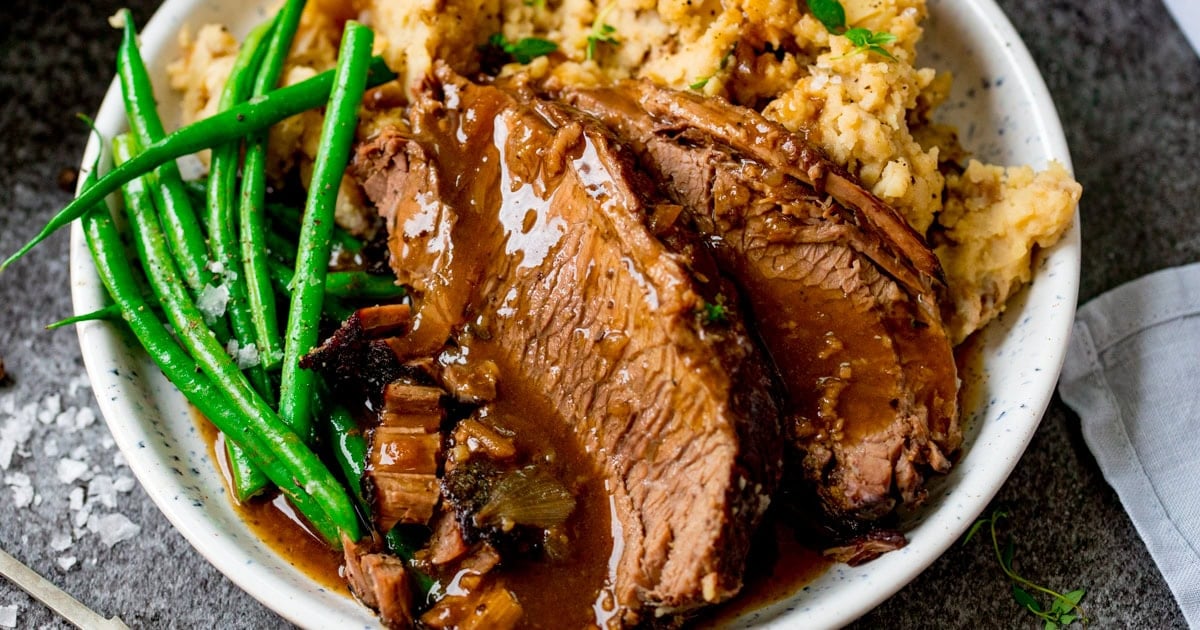 A plate of slow cooker roast beef with brown gravy, mashed potato, and green beans, served on a white plate.