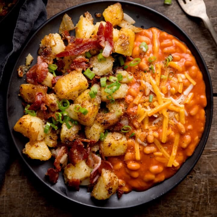 A black plate with baked beans topped with grated cheese on one side and a hearty potato and bacon hash with potatoes, bacon pieces, and chopped spring onions on the other.