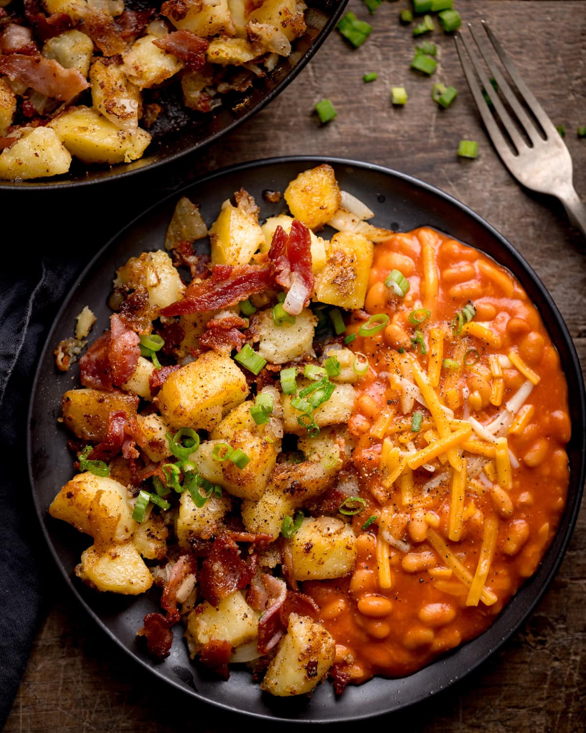 A black plate with potato and bacon hash, roast potatoes, and chopped spring onions on one side, and baked beans topped with grated cheddar cheese on the other. A fork and extra garnish are nearby.