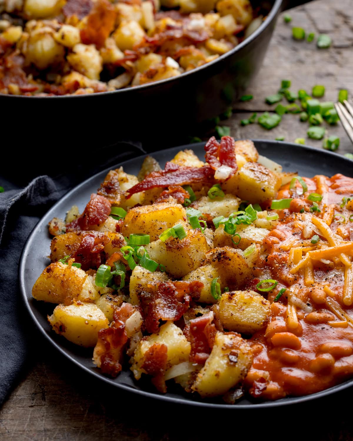 A plate of seasoned potato and bacon hash with crispy bacon and chopped spring onions, served with baked beans topped with grated cheese.