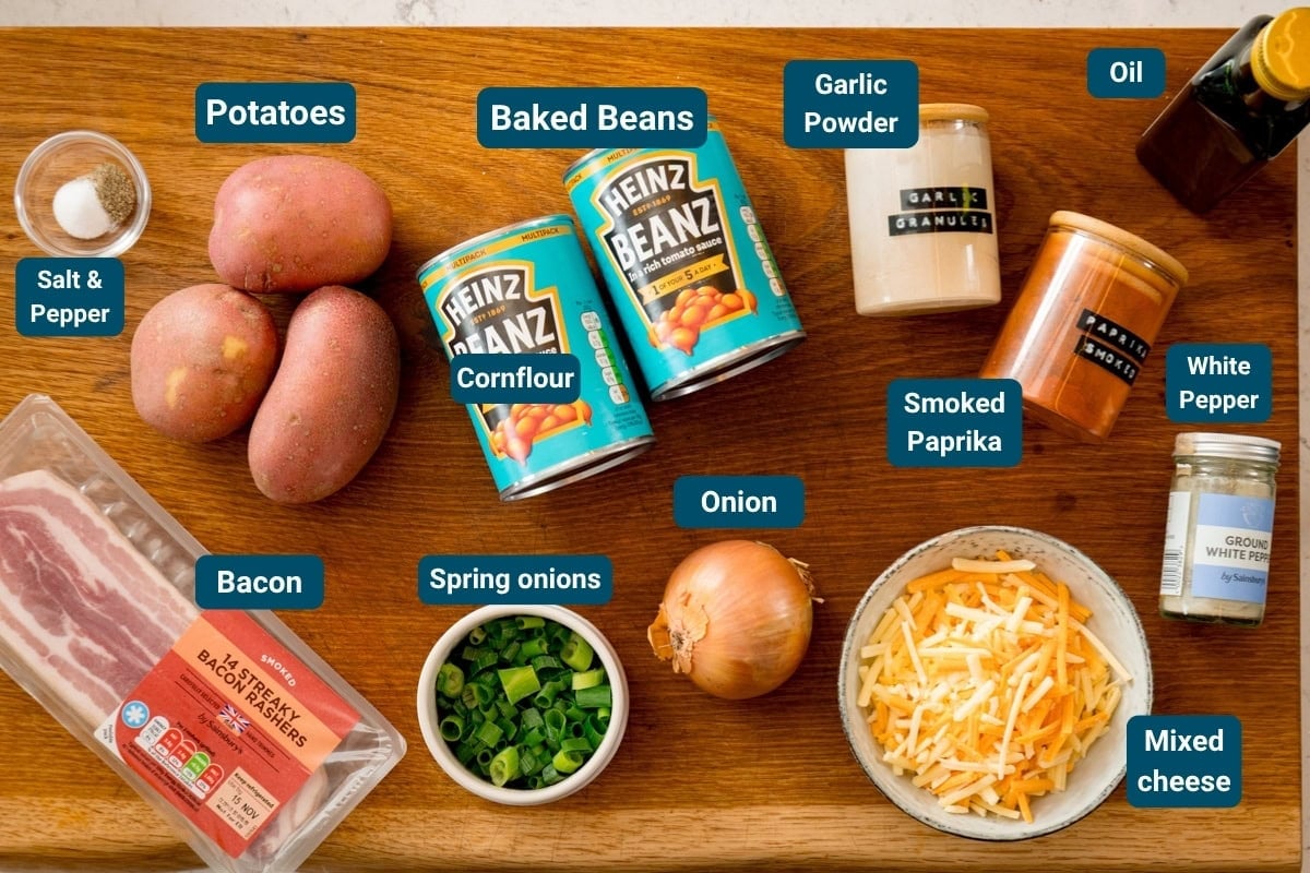 A wooden surface with labelled ingredients: potatoes, baked beans, cornflour, spring onions, onion, bacon, mixed cheese, smoked paprika, white pepper, garlic powder, oil, salt and pepper.