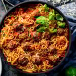 Spaghetti and meatballs in a black pan, topped with grated cheese and fresh basil, served with rich sauce.