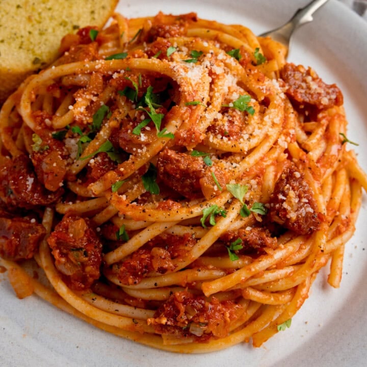 Close up of a white plate of bucatini with tomato chorizo sauce and grated cheese, garnished with parsley, served with garlic bread.
