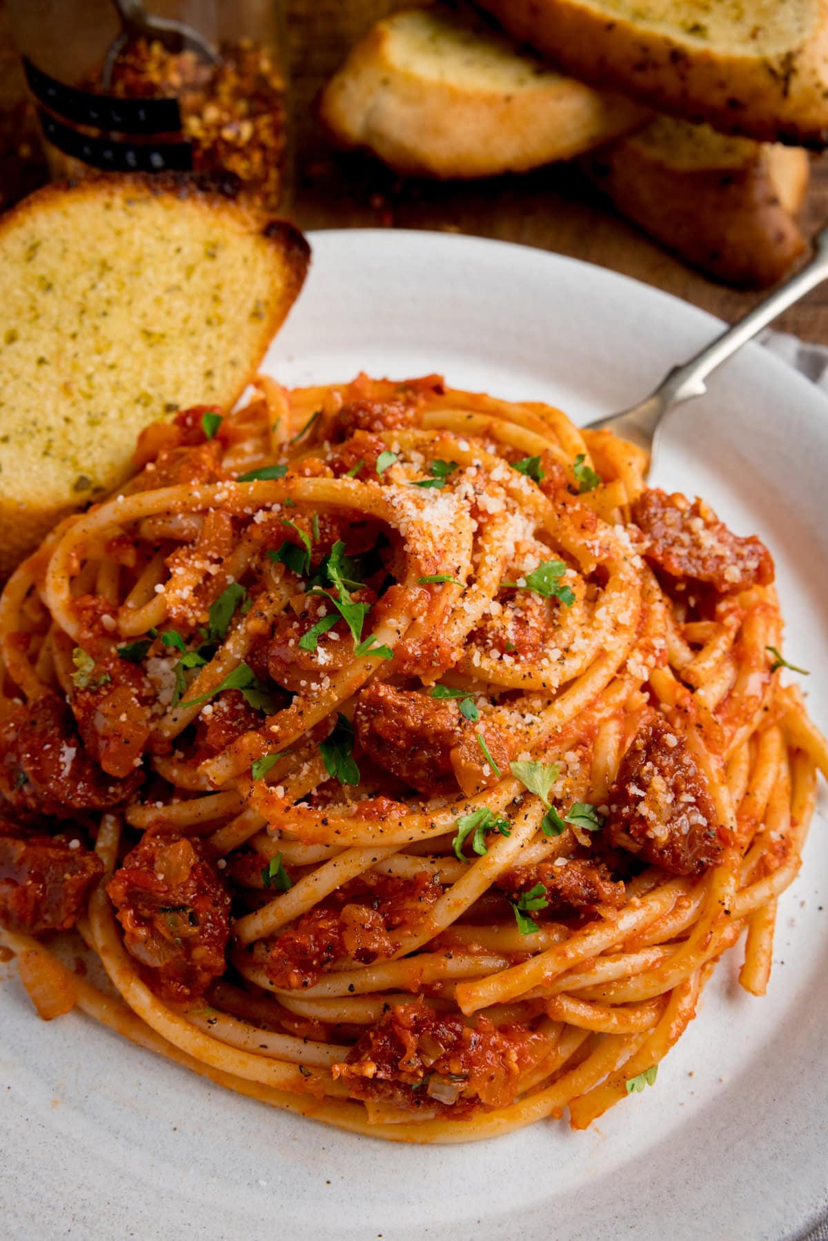 A plate of chorizo bucatini pasta with spicy tomato sauce is served with a slice of garlic bread. There is more garlic bread inthe background.