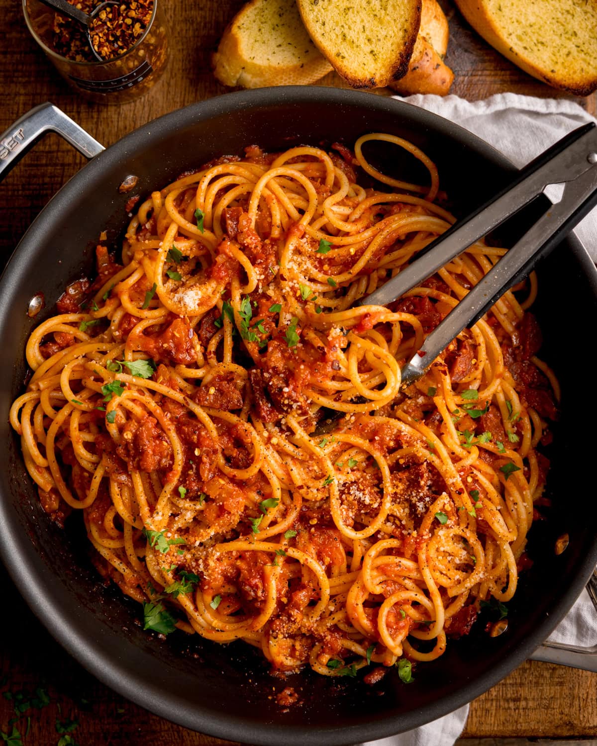 A frying pan of chorizo pasta with tomato sauce, topped with grated cheese and parsley. A set of tongs is grabbing some pasta. There are garlic bread slices, and red chilli flakes nearby.