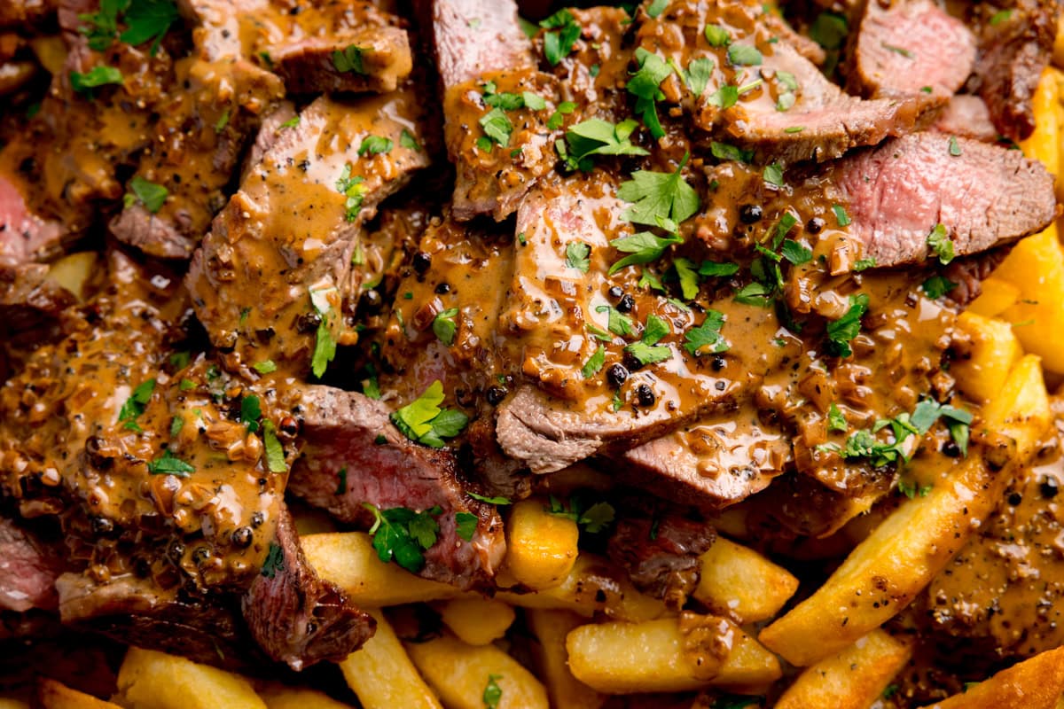 Close up wide image of sliced steak with Peppercorn Sauce and chopped parsley served over a bed of oven chips.