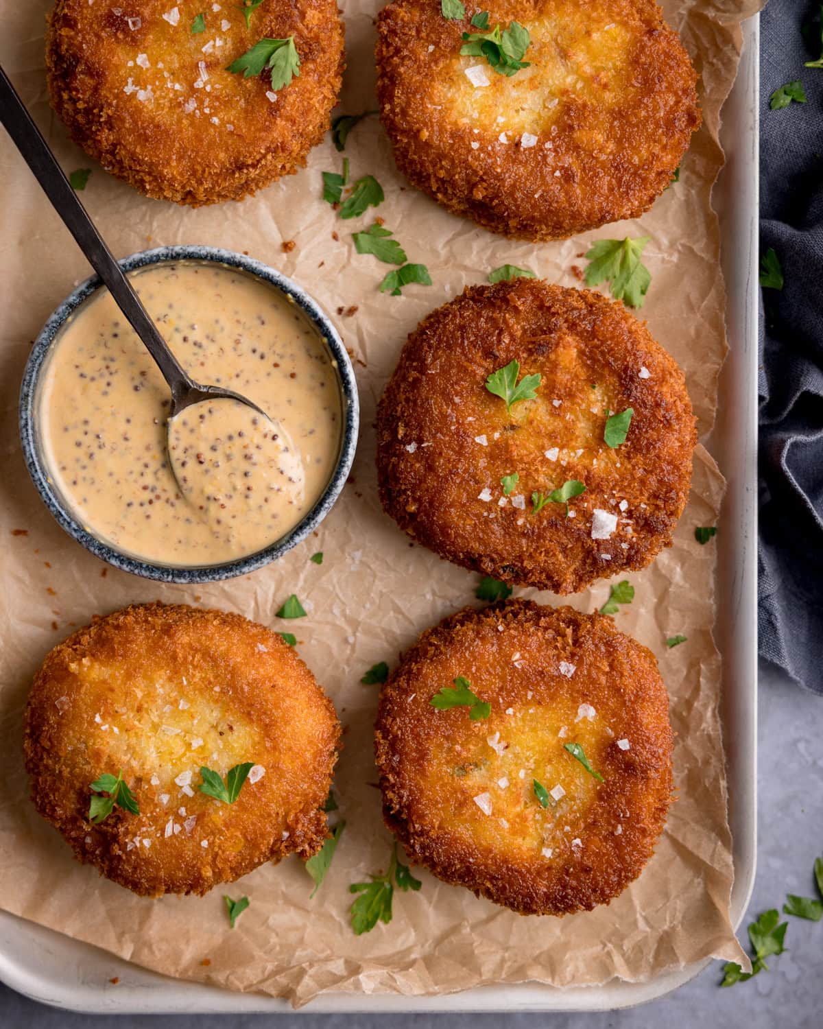 Five breaded and fried tuna fishcakes on parchment paper, garnished with parsley, served with a small bowl of wholegrain mustard dip and a spoon.