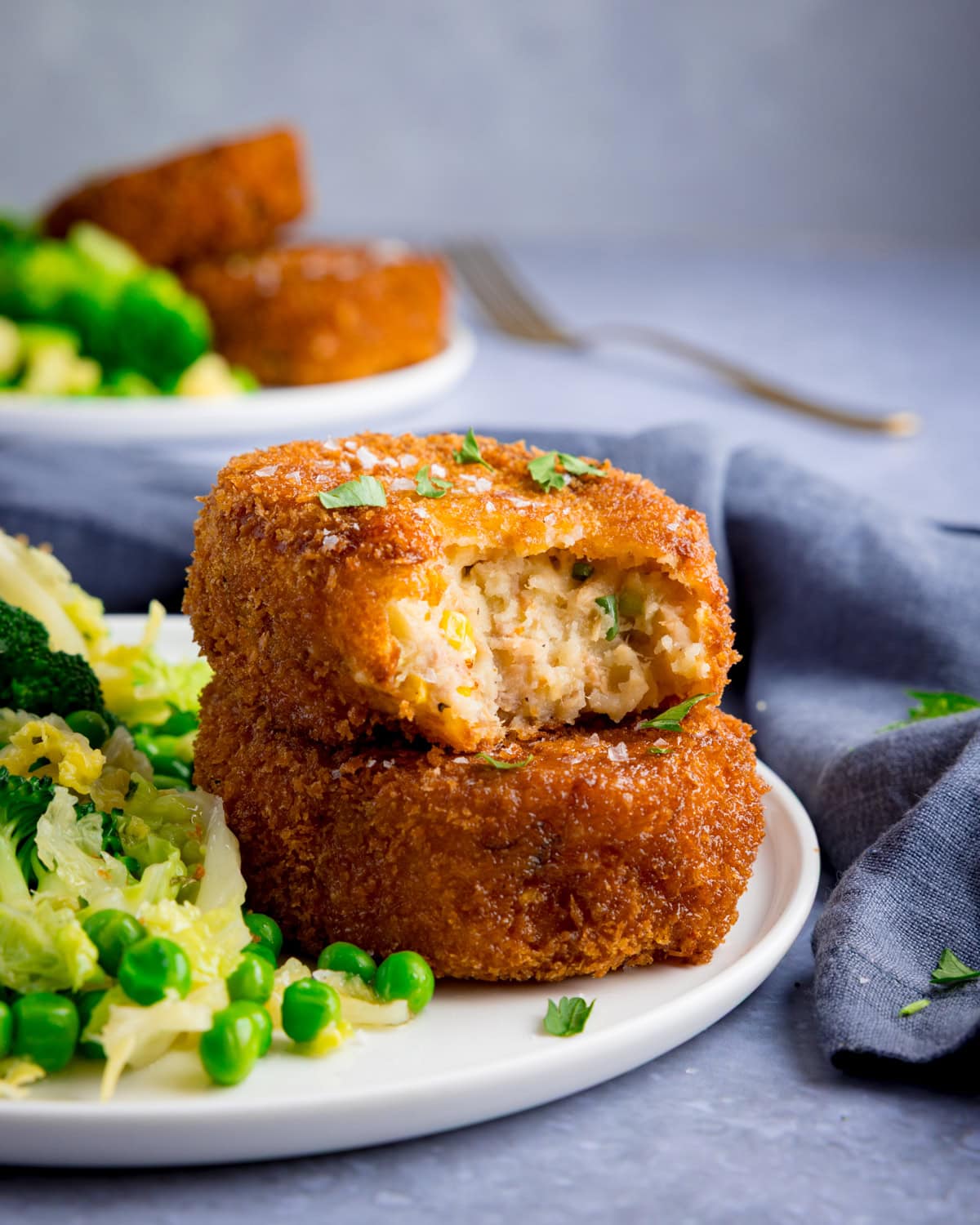Two breaded tuna fish cakes are stacked on a plate, one partially eaten, served with peas, broccoli, and shredded cabbage.