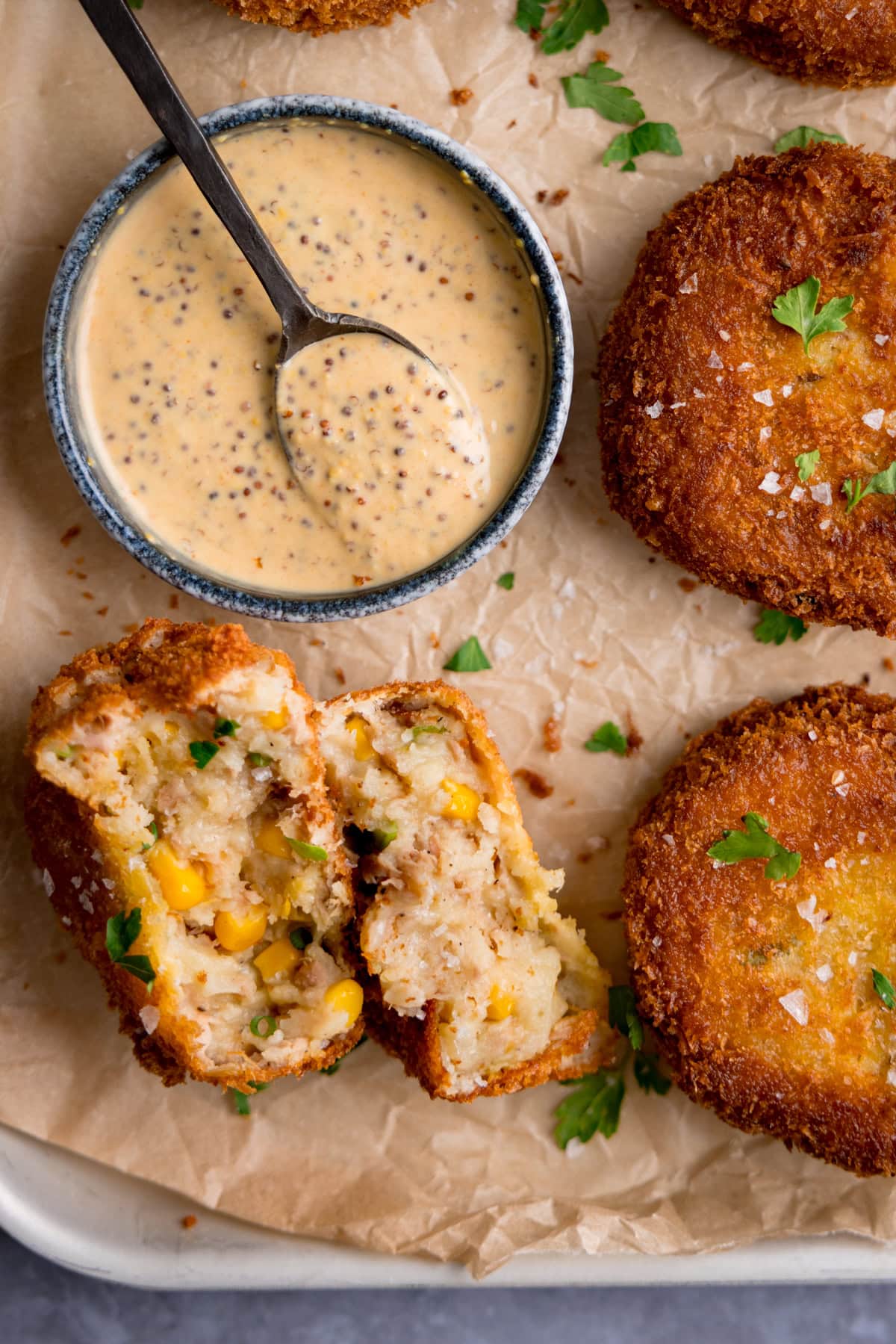 Four golden-brown tuna fish cakes rest on baking parchment beside a small bowl of creamy sauce; one is cut open, showing sweetcorn and fresh herbs inside.