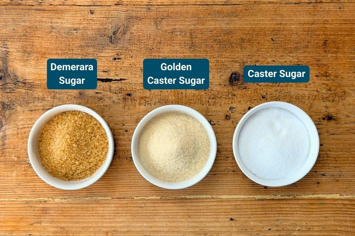 Three small bowls on a wooden surface, each holding Demerara Sugar, Golden Caster Sugar, and Caster Sugar, labelled accordingly.