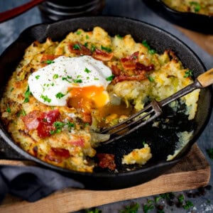 A cast iron frying pan containing bubble and squeak with bacon, topped with a poached egg, garnished with chopped parsley; a fork rests inside, and the yolk is runny.