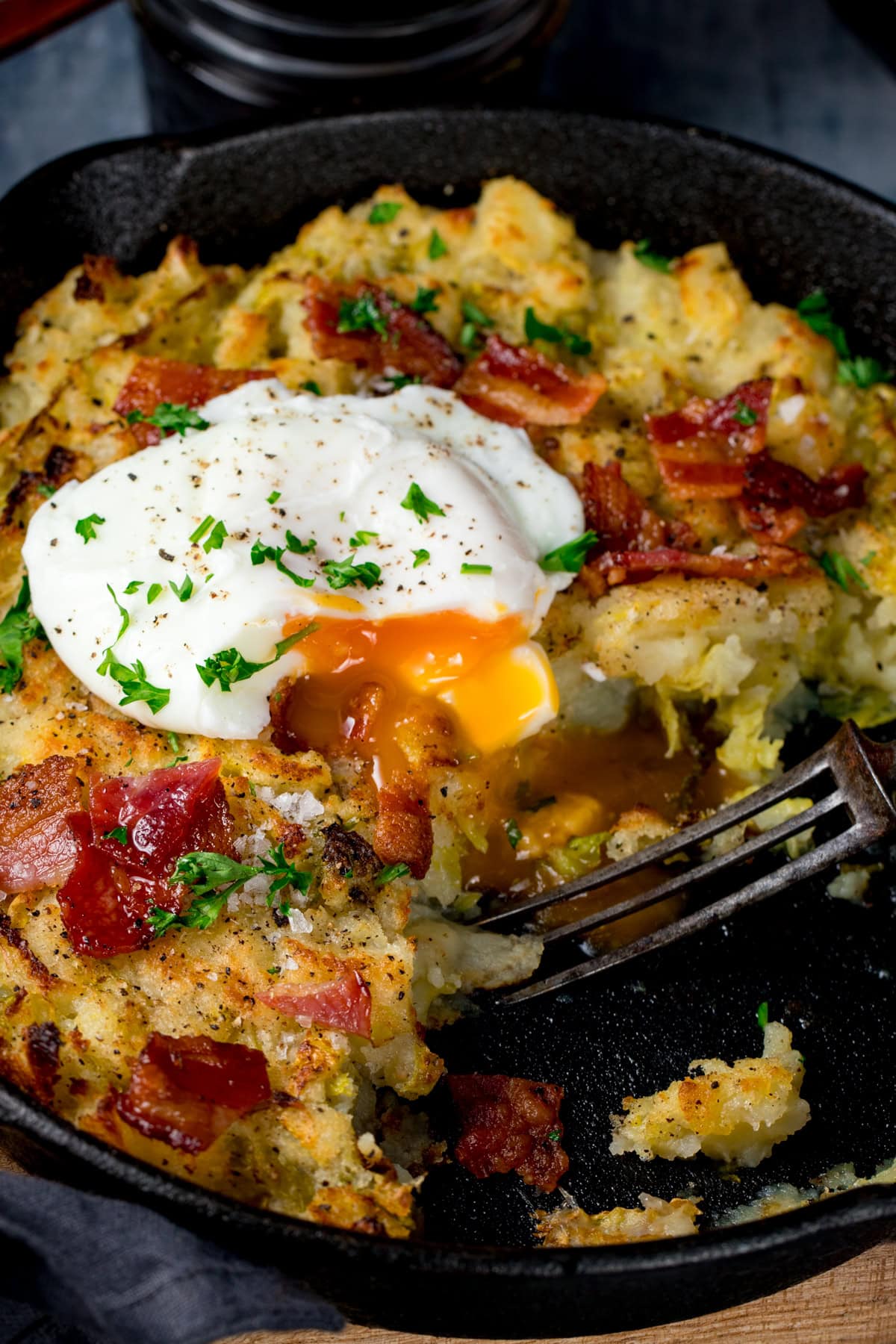 A frying pan with bubble and squeak with bacon, a poached egg with a runny yolk, and chopped parsley, with a fork inside.