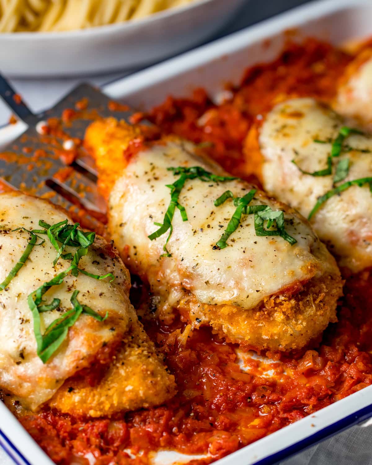 Breaded chicken parmesan breasts topped with melted cheese and fresh basil, on tomato sauce in a baking dish, pasta in background.