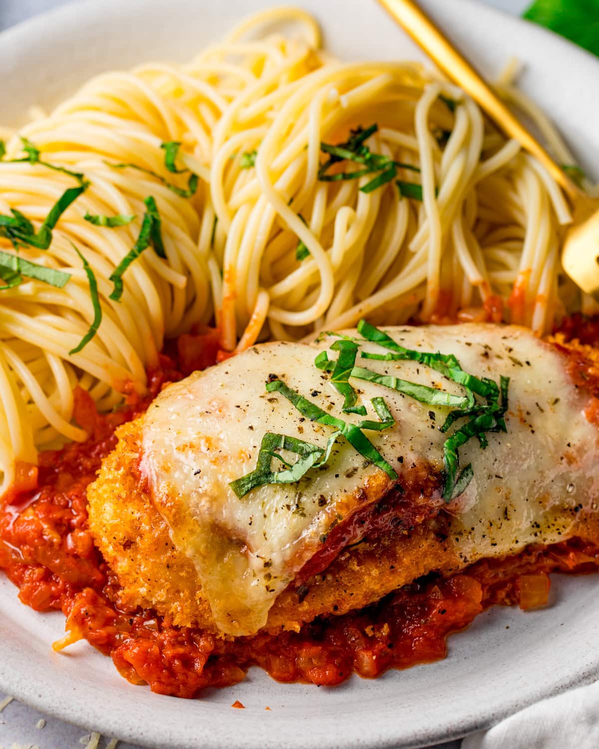 Plate of breaded chicken topped with melted cheese and basil, served on tomato sauce beside spaghetti garnished with chopped basil.