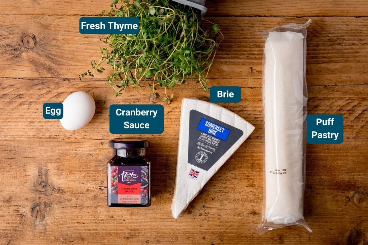 Labelled ingredients for cranberry brie bites, including an egg, a jar of cranberry sauce, a wedge of brie, puff pastry, and fresh thyme on a wooden surface.