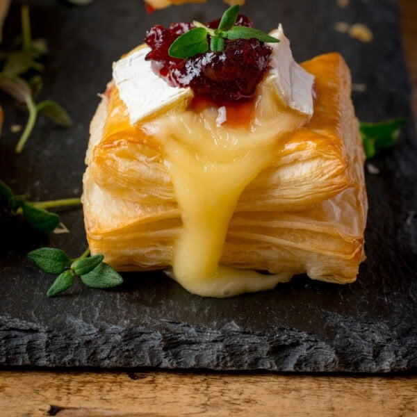 Close up of a square puff pastry topped with melted brie cheese and cranberry sauce, garnished with fresh thyme, sitting on a dark slate serving board.