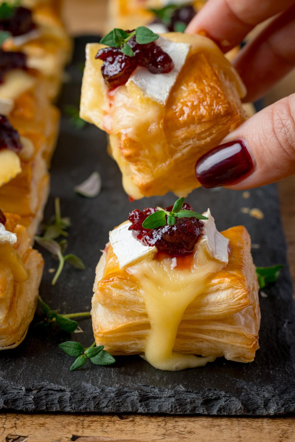 A hand picks up a Cranberry and Brie Bite topped with melted brie, cranberry sauce and fresh thyme. Further cranberry brie bites are on a slate serving board underneath.