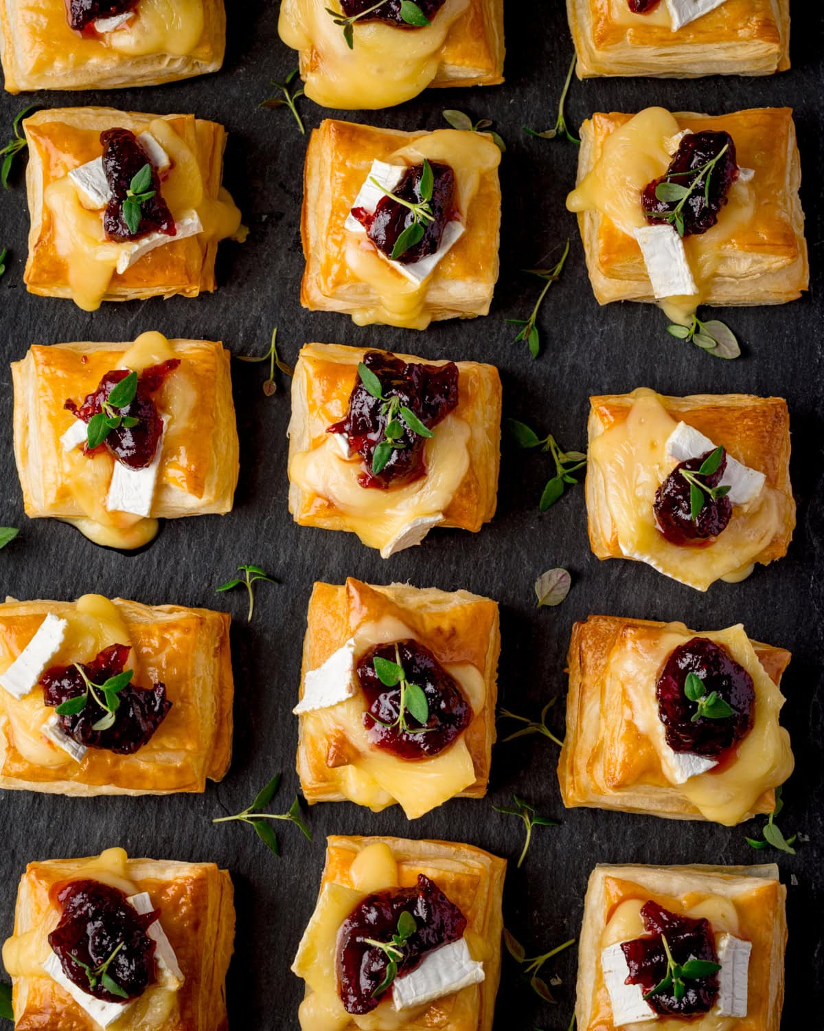 Overhead image of squares of puff pastry topped with melted brie cheese, cranberry sauce, and fresh thyme arranged on a dark slate board in rows.