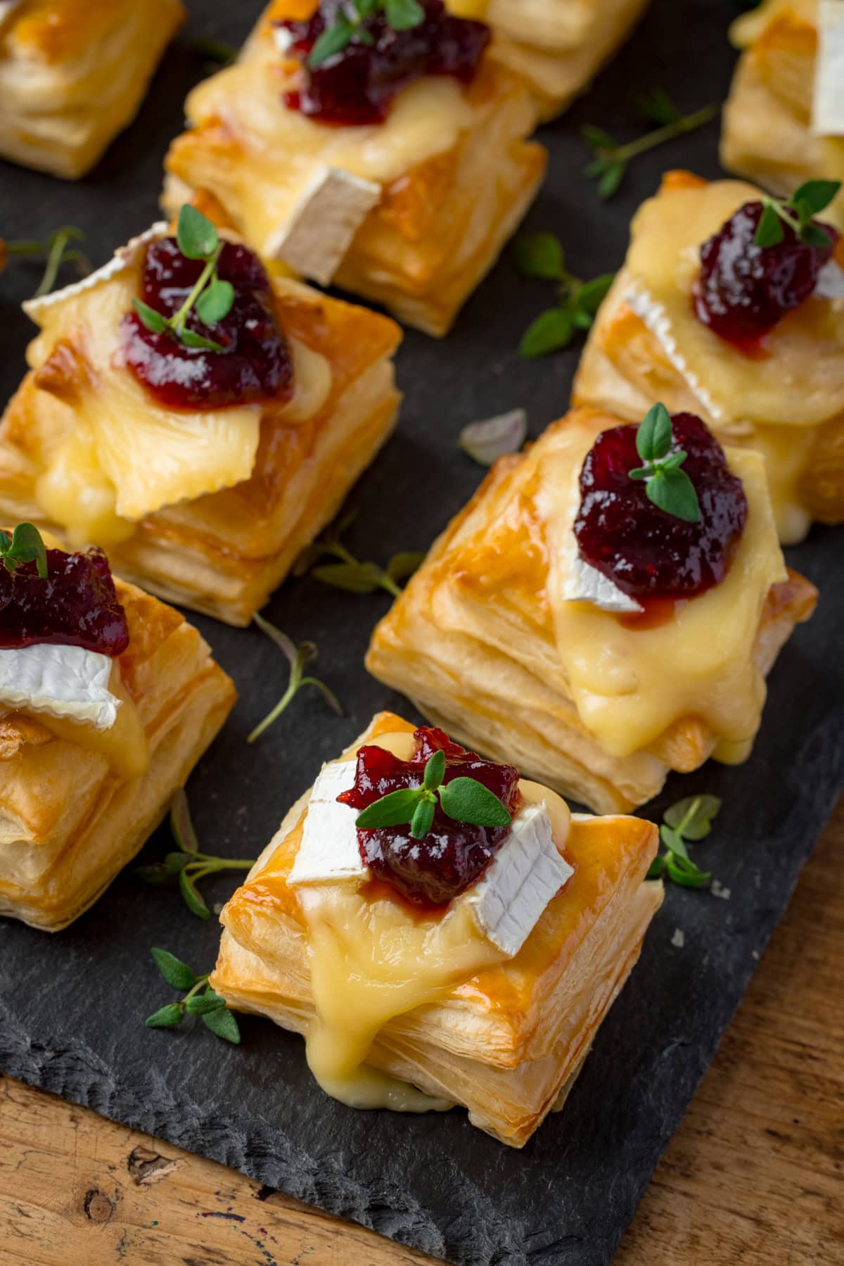 Small puff pastry squares topped with melted brie, cranberry sauce, and fresh thyme, arranged on a dark slate serving board.