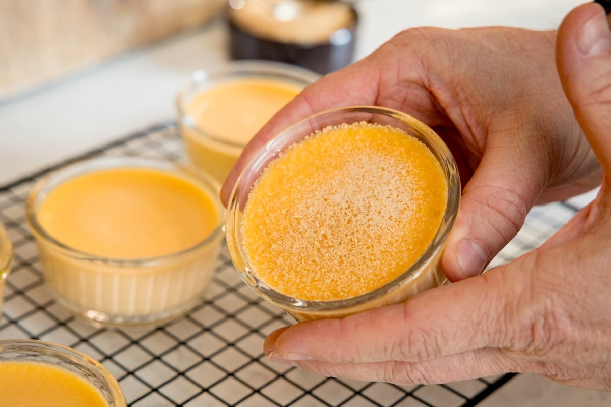 A person holds a small glass dish of custard with sugar sprinkled on top, with more custard dishes cooling on a wire rack in the background.
