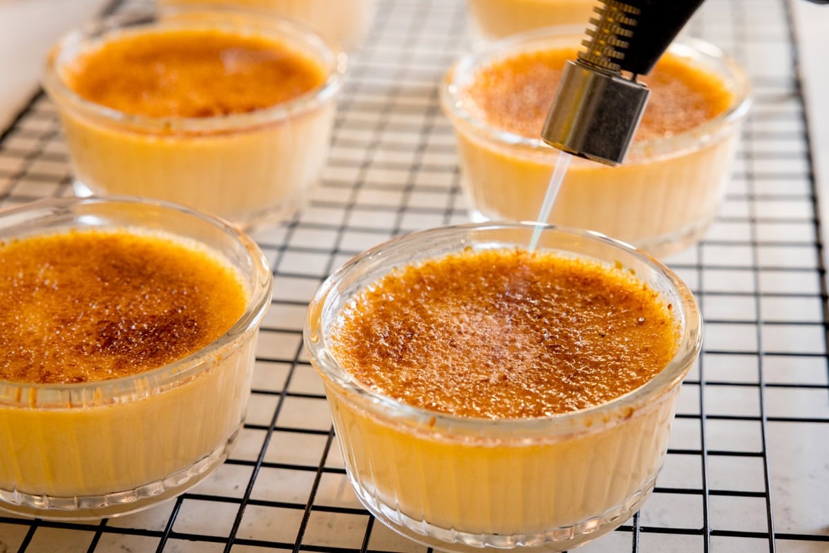 Several ramekins of crème brûlée are shown, with a kitchen blowtorch caramelising the sugar on top of one, all on a cooling rack.