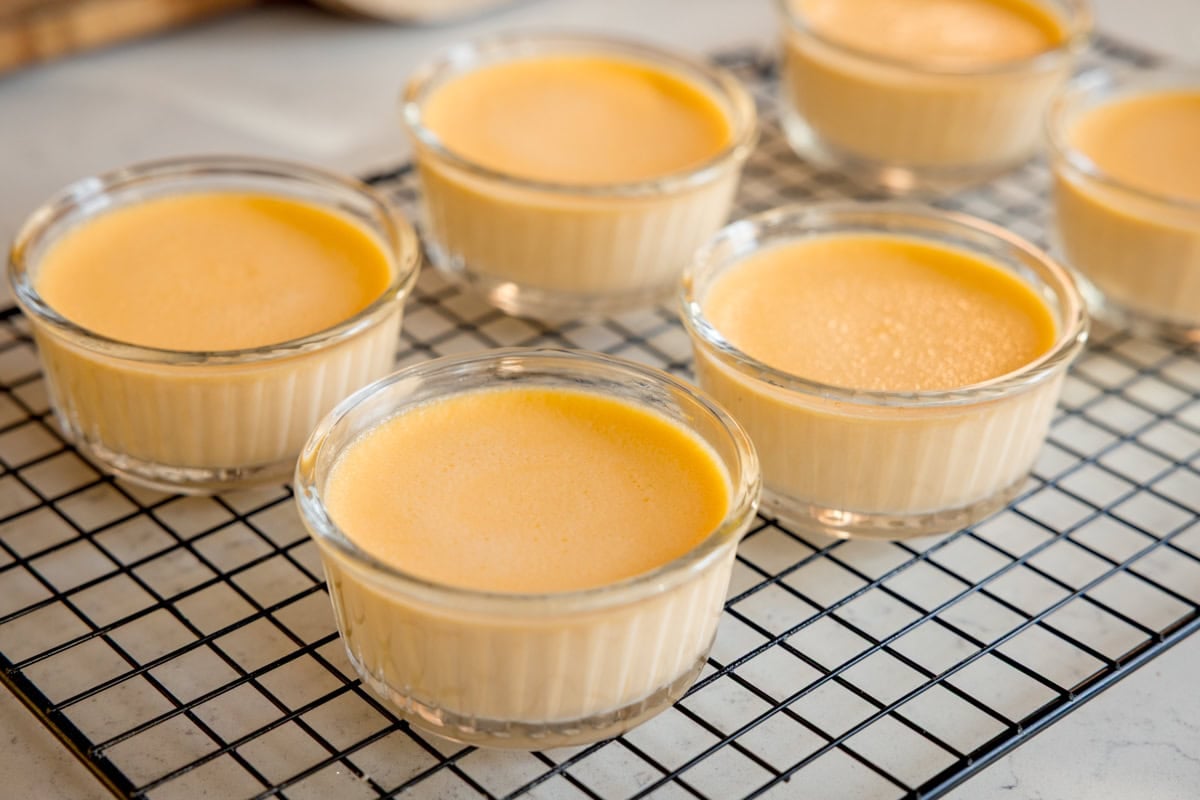 Six glass ramekins filled with smooth, yellow custard are cooling on a black wire rack set on a light-coloured worktop.