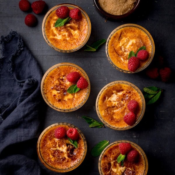 Six ramekins of crème brûlée topped with fresh raspberries and mint leaves are arranged on a dark surface, with a bowl of brown sugar and a dark serviette nearby.
