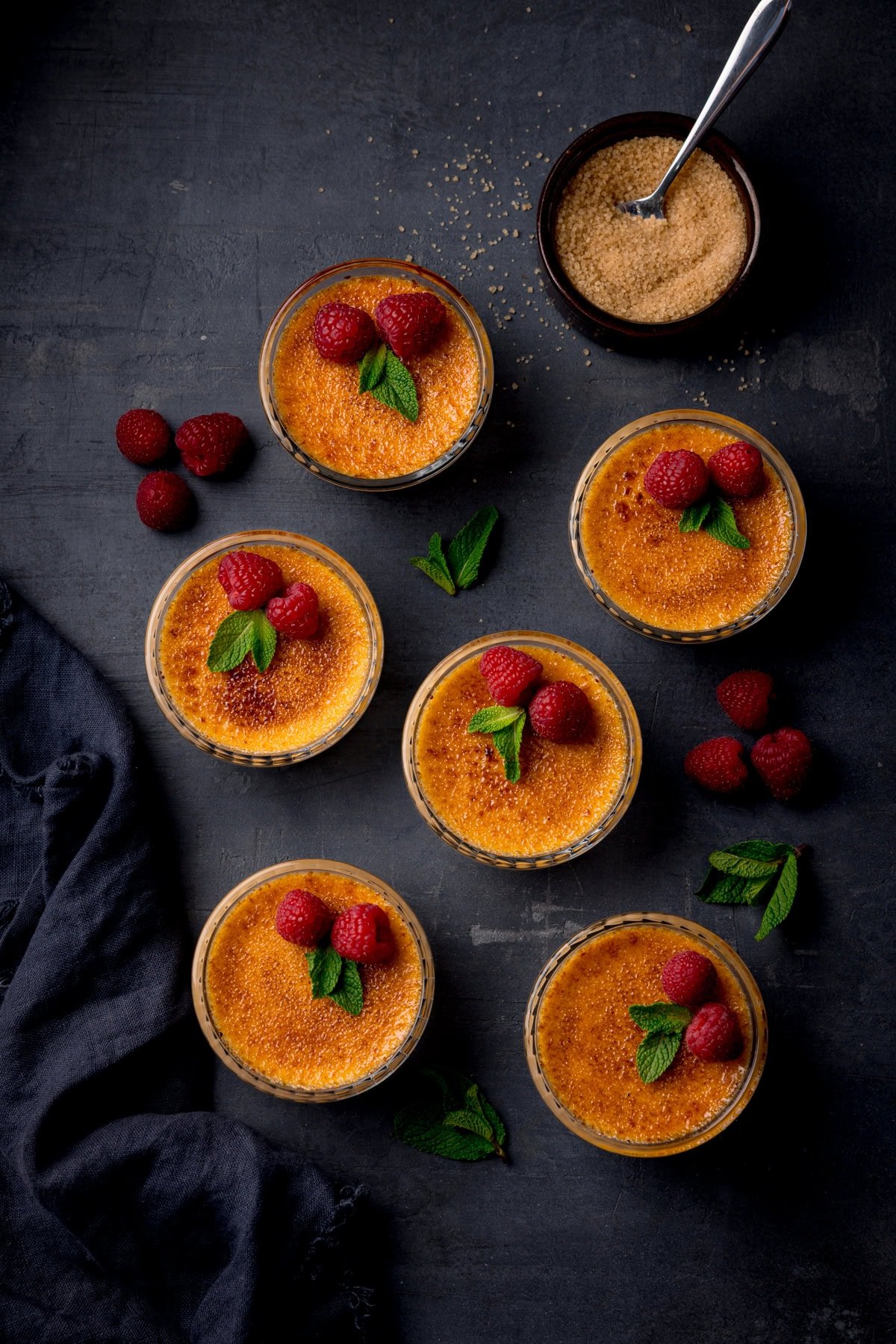 Six ramekins of crème brûlée topped with raspberries and mint, surrounded by loose raspberries, mint leaves, a bowl of brown sugar, and a dark cloth on a dark surface.