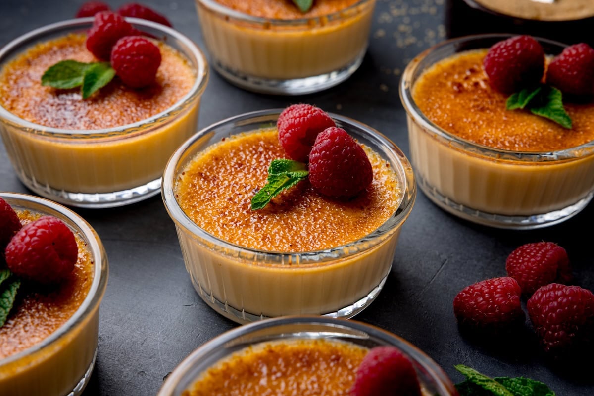 Small glass ramekins of crème brûlée are topped with raspberries and mint leaves, arranged on a dark surface.