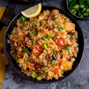 A black bowl filled with Ham and Pineapple Fried Rice with diced vegetables, pineapple chunks, sliced spring onions, and a lemon wedge. The bowl is on a dark grey surface.