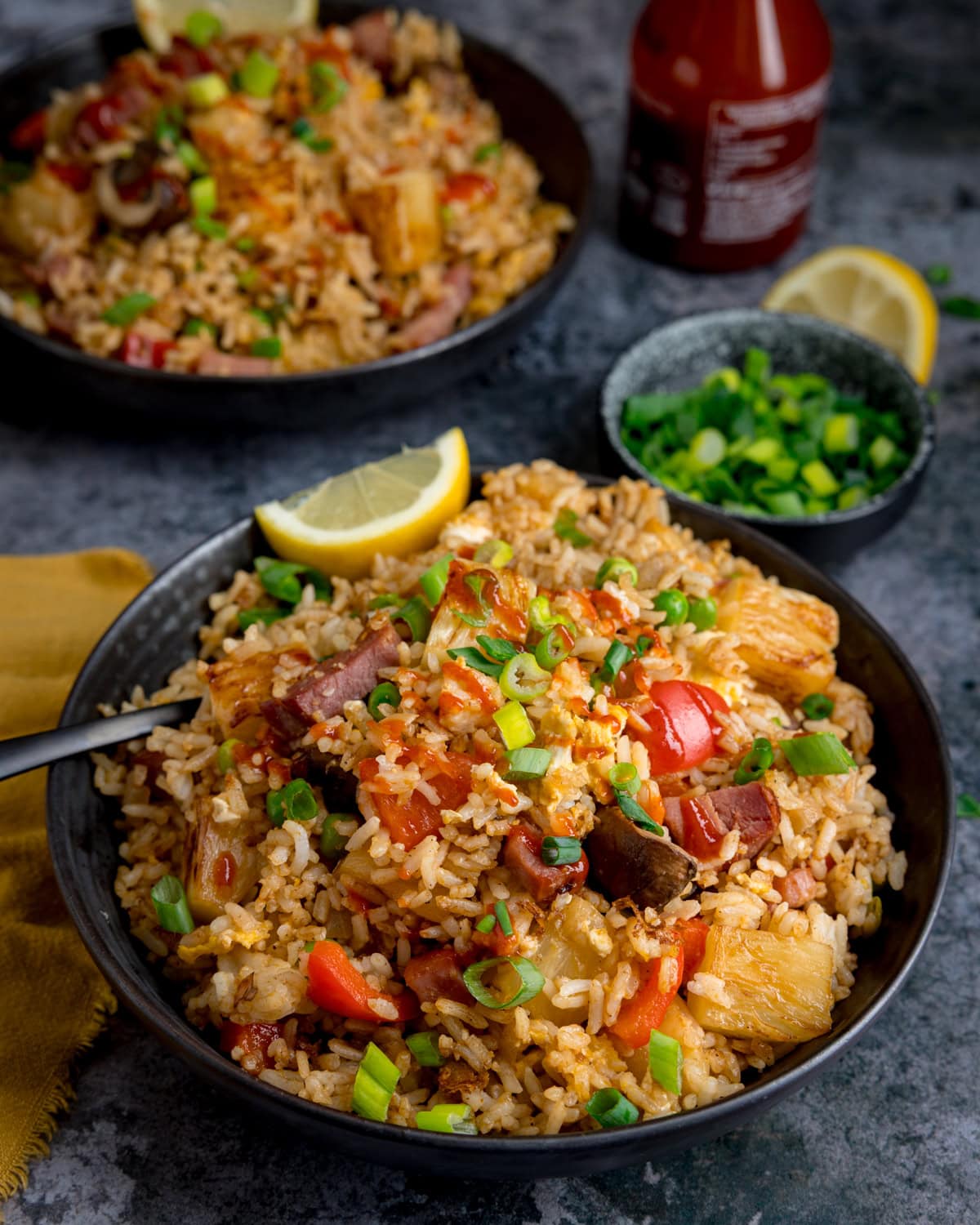 A bowl of ham and pineapple fried rice with chunks of ham, pineapple, pepper and mushroom, topped with chopped spring onions and lemon wedges, a fork is resting in the bowl. There is a further bowl of rice in the background alongside a small dish of spring onions.