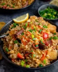 A bowl of ham and pineapple fried rice with chunks of ham, pineapple, pepper and mushroom, topped with chopped spring onions and a lemon wedge. There is a further bowl of rice in the background alongside a small dish of spring onions.