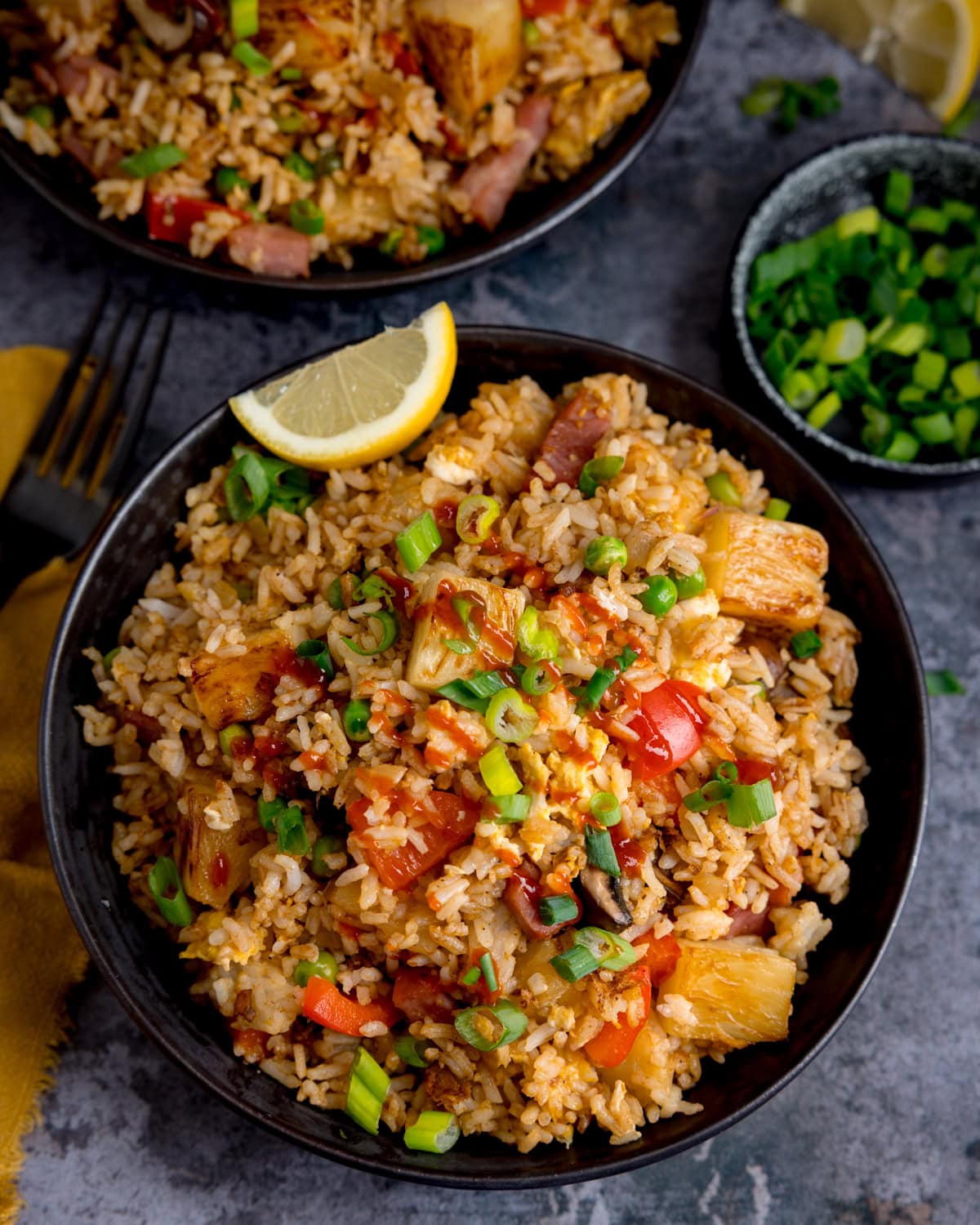 Overhead image of a bowl of Ham and Pineapple Fried Rice with chopped spring onions, diced vegetables, pineapple, and a lemon wedge on top, served in a black bowl on a grey surface next to a small dish of spring onions and a further bowl of fried rice.